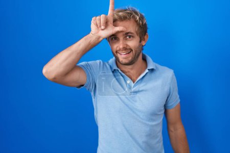 Photo for Caucasian man standing over blue background making fun of people with fingers on forehead doing loser gesture mocking and insulting. - Royalty Free Image