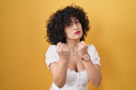Photo for Young brunette woman with curly hair standing over yellow background ready to fight with fist defense gesture, angry and upset face, afraid of problem - Royalty Free Image