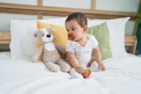 Foto de Adorable bebé hispano chupando chupete sentado en la cama en el dormitorio - Imagen libre de derechos