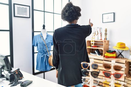 Foto de Mujer joven africana trabajando como gerente en boutique minorista posando hacia atrás apuntando hacia adelante con la mano del dedo - Imagen libre de derechos