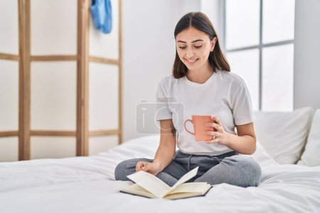 Foto de Mujer hispana joven bebiendo taza de libro de lectura de café en el dormitorio - Imagen libre de derechos