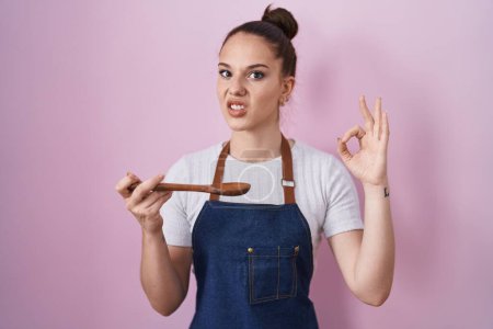 Photo for Young hispanic girl wearing professional cook apron holding wood spoon clueless and confused expression. doubt concept. - Royalty Free Image