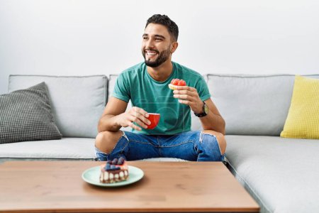 Téléchargez les photos : Jeune homme arabe prenant le petit déjeuner assis sur le canapé à la maison - en image libre de droit