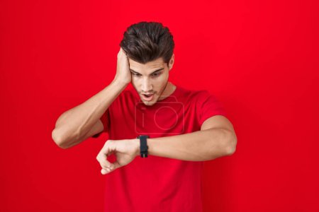 Foto de Joven hombre hispano de pie sobre fondo rojo mirando la hora del reloj preocupado, con miedo de llegar tarde - Imagen libre de derechos
