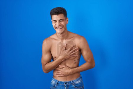 Foto de Young hispanic man standing shirtless over blue background smiling and laughing hard out loud because funny crazy joke with hands on body. - Imagen libre de derechos