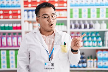 Téléchargez les photos : Jeune homme chinois travaillant à la pharmacie pharmacie surpris pointant du doigt sur le côté, bouche ouverte expression étonnée. - en image libre de droit