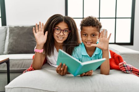Photo for Two siblings lying on the sofa reading a book waiving saying hello happy and smiling, friendly welcome gesture - Royalty Free Image