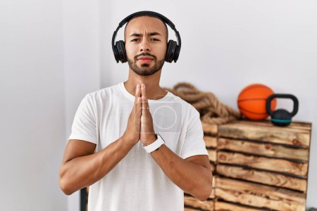 Foto de Hombre afroamericano escuchando música usando auriculares en el gimnasio mendigando y rezando con las manos juntas con la expresión de esperanza en la cara muy emocional y preocupado. mendicidad. - Imagen libre de derechos