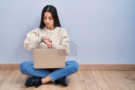 Foto de Mujer joven usando el ordenador portátil sentado en el suelo en casa comprobando la hora en el reloj de pulsera, relajado y seguro - Imagen libre de derechos