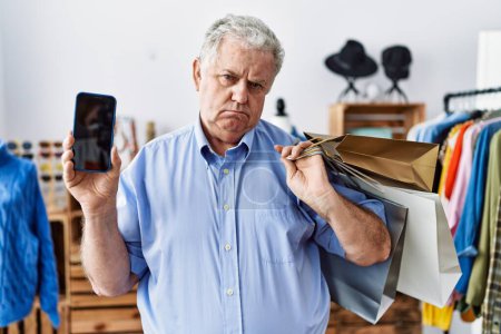 Foto de Senior man with grey hair holding shopping bags and showing smartphone screen depressed and worry for distress, crying angry and afraid. sad expression. - Imagen libre de derechos
