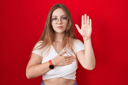Téléchargez les photos : Jeune femme caucasienne debout sur fond rouge jurant avec la main sur la poitrine et la paume ouverte, faire un serment de fidélité promesse - en image libre de droit