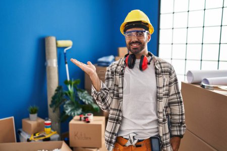 Téléchargez les photos : Jeune homme hispanique avec barbe travaillant à la rénovation de la maison souriant gai présentation et pointant avec la paume de la main regardant la caméra. - en image libre de droit