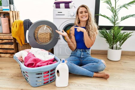 Téléchargez les photos : Young beautiful woman doing laundry sitting by wicker basket amazed and smiling to the camera while presenting with hand and pointing with finger. - en image libre de droit