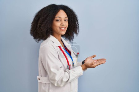 Téléchargez les photos : Jeune femme afro-américaine portant l'uniforme de médecin et stéthoscope pointant de côté avec les mains ouvertes paumes montrant l'espace de copie, présentant la publicité souriant heureux excité - en image libre de droit