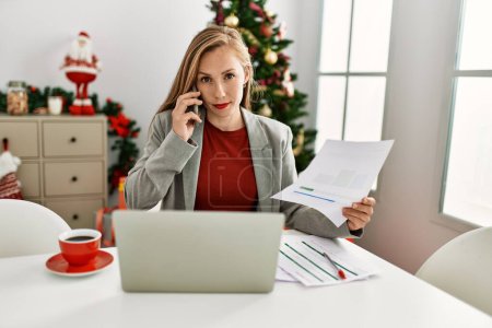 Foto de Young caucasian woman talking on the smartphone working sitting by christmas tree at home - Imagen libre de derechos