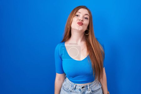 Foto de Redhead woman standing over blue background looking at the camera blowing a kiss on air being lovely and sexy. love expression. - Imagen libre de derechos