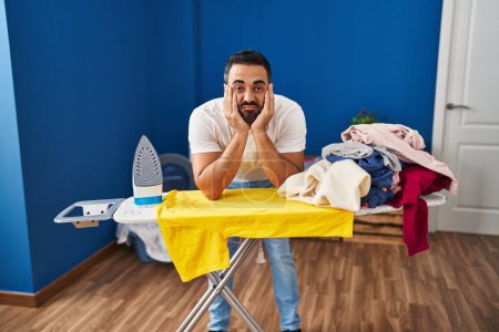 Foto de Joven hispano cansado apoyado en tabla de planchar en la lavandería - Imagen libre de derechos