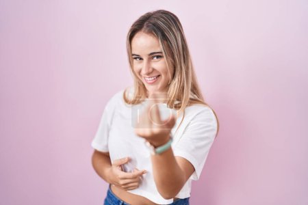 Foto de Joven mujer rubia de pie sobre fondo rosa haciendo señas ven aquí gesto con la mano invitando a la bienvenida feliz y sonriente - Imagen libre de derechos