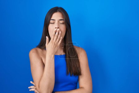 Photo for Hispanic woman standing over blue background bored yawning tired covering mouth with hand. restless and sleepiness. - Royalty Free Image