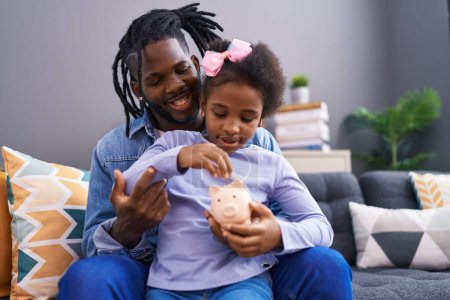 Photo for Father and daughter inserting coin on piggy bank sitting on sofa at home - Royalty Free Image