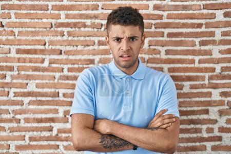 Photo for Brazilian young man standing over brick wall skeptic and nervous, disapproving expression on face with crossed arms. negative person. - Royalty Free Image