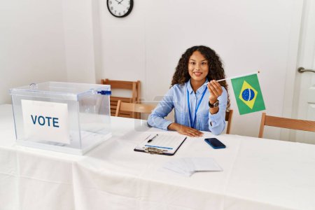 Foto de Joven latina sonriendo confiada sosteniendo bandera brasileña trabajando en colegio electoral - Imagen libre de derechos