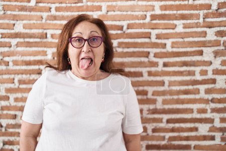 Téléchargez les photos : Femme âgée avec des lunettes debout sur le mur de briques collant la langue heureux avec une expression drôle. concept d'émotion. - en image libre de droit