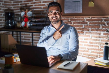 Foto de Hispanic man with beard working at the office at night cheerful with a smile on face pointing with hand and finger up to the side with happy and natural expression - Imagen libre de derechos