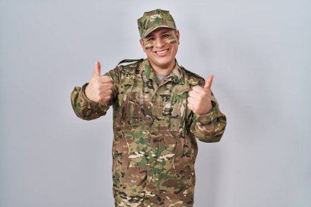 Foto de Hispanic young man wearing camouflage army uniform success sign doing positive gesture with hand, thumbs up smiling and happy. cheerful expression and winner gesture. - Imagen libre de derechos