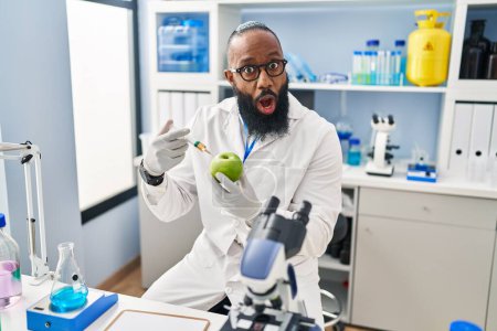 Téléchargez les photos : African american man working at scientist laboratory with apple afraid and shocked with surprise and amazed expression, fear and excited face. - en image libre de droit