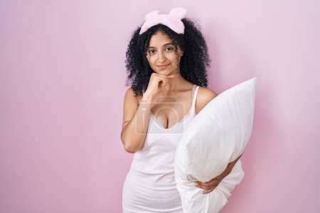Téléchargez les photos : Hispanic woman with curly hair wearing sleep mask and pajama holding pillow looking confident at the camera smiling with crossed arms and hand raised on chin. thinking positive. - en image libre de droit