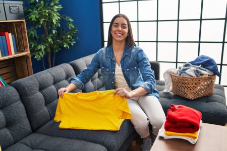 Photo for Young beautiful hispanic woman folding clothes sitting on sofa at home - Royalty Free Image