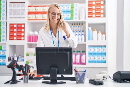 Photo for Young blonde woman pharmacist talking on smartphone using computer at pharmacy - Royalty Free Image