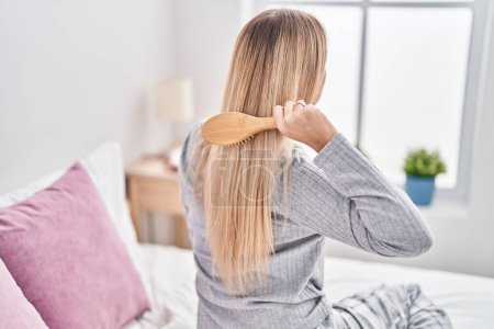 Téléchargez les photos : Jeune femme peigner les cheveux assis sur le lit dans la chambre - en image libre de droit