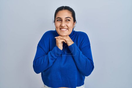 Foto de Young hispanic woman standing over isolated background laughing nervous and excited with hands on chin looking to the side - Imagen libre de derechos