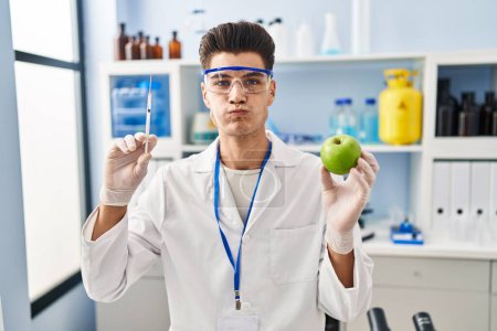 Téléchargez les photos : Young hispanic man working at scientist laboratory holding apple puffing cheeks with funny face. mouth inflated with air, catching air. - en image libre de droit