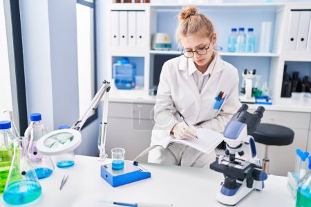 Photo for Young blonde woman scientist writing on notebook weighing liquid at laboratory - Royalty Free Image