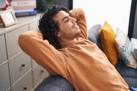 Foto de Young latin man relaxed with hands on head sitting on sofa at home - Imagen libre de derechos