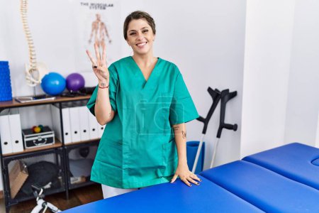 Téléchargez les photos : Young hispanic woman wearing physiotherapist uniform standing at clinic showing and pointing up with fingers number three while smiling confident and happy. - en image libre de droit