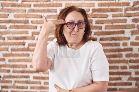 Foto de Mujer mayor con gafas de pie sobre ladrillos pared disparando y matándose señalando la mano y los dedos a la cabeza como arma, gesto suicida. - Imagen libre de derechos