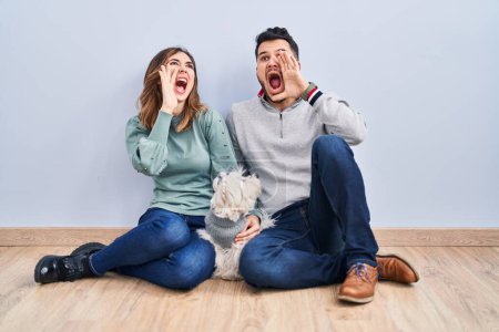 Téléchargez les photos : Young hispanic couple sitting on the floor with dog shouting and screaming loud to side with hand on mouth. communication concept. - en image libre de droit