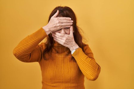 Foto de Middle age hispanic woman standing over yellow background covering eyes and mouth with hands, surprised and shocked. hiding emotion - Imagen libre de derechos