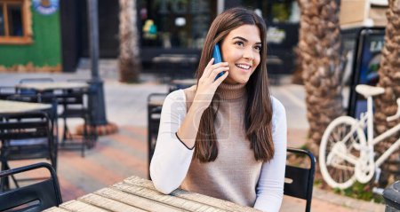 Téléchargez les photos : Jeune belle femme hispanique parlant sur le smartphone assis sur la table à la terrasse du café - en image libre de droit