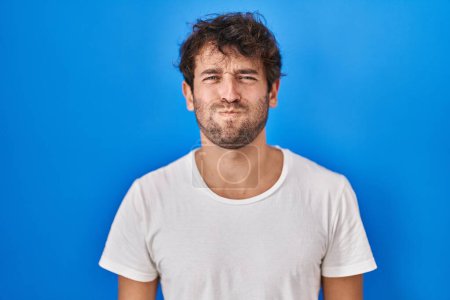 Téléchargez les photos : Hispanic young man standing over blue background puffing cheeks with funny face. mouth inflated with air, crazy expression. - en image libre de droit