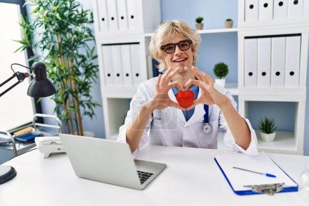 Photo for Young blond man doctor smiling confident holding heart at clinic - Royalty Free Image