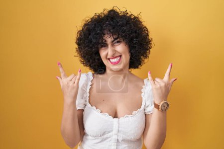 Foto de Young brunette woman with curly hair standing over yellow background shouting with crazy expression doing rock symbol with hands up. music star. heavy music concept. - Imagen libre de derechos