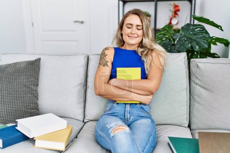 Photo for Young woman sitting on sofa hugging book at home - Royalty Free Image