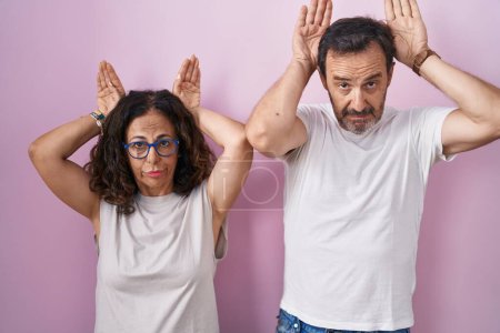 Photo for Middle age hispanic couple together over pink background doing bunny ears gesture with hands palms looking cynical and skeptical. easter rabbit concept. - Royalty Free Image