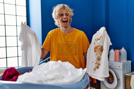 Photo for Young man holding clean white t shirt and t shirt with dirty stain smiling and laughing hard out loud because funny crazy joke. - Royalty Free Image