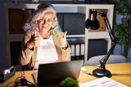 Foto de Middle age woman with grey hair working using computer laptop late at night success sign doing positive gesture with hand, thumbs up smiling and happy. cheerful expression and winner gesture. - Imagen libre de derechos
